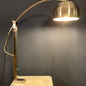Puede incluir: Una lámpara de escritorio de plata arqueada con una base de mármol blanco. La lámpara tiene una pantalla redonda de plata con un diseño acanalado.