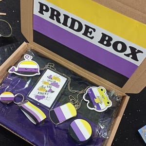 Może przedstawiać: Tekturowe pudełko z nadrukiem "PRIDE BOX" na pokrywie. Pudełko jest otwarte i zawiera różne przedmioty, w tym czarną koszulkę z białym wzorem wieloryba, brelok do kluczy z żółto-biało-fioletową wstążką, naszyjnik z żółto-biało-fioletowym znacznikiem i naklejkę z motywem żaby. Pudełko jest wyłożone fioletowym materiałem.