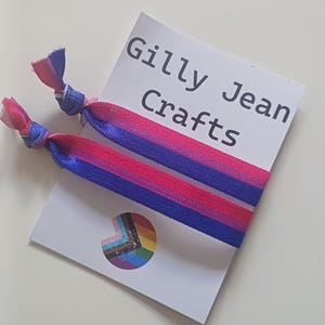 May include: Two hair ties with a pink, purple, and blue striped pattern, knotted at the ends. They rest on a white card with the text "Gilly Jean Crafts" and a rainbow pride symbol.