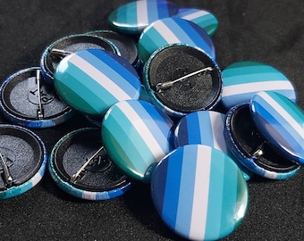 Puede incluir: Primer plano de un montón de botones de solapa rayados en azul, verde y blanco. Los botones tienen un cierre de seguridad plateado y están dispuestos en una pila.