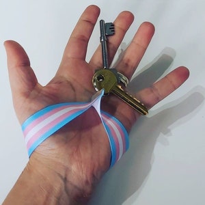Puede incluir: Un juego de llaves con una cinta con los colores de la bandera del orgullo transg&eacute;nero: azul claro, rosa y blanco. Las llaves se sostienen en una mano sobre un fondo blanco. Una llave es plateada y la otra dorada.