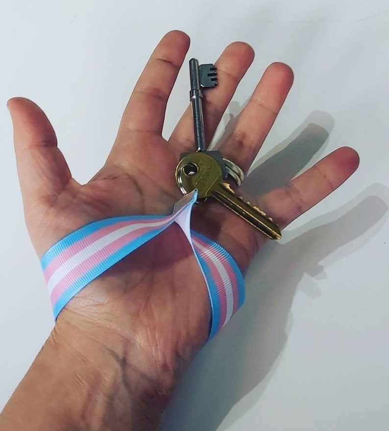 Puede incluir: Un juego de llaves con una cinta con los colores de la bandera del orgullo transg&eacute;nero: azul claro, rosa y blanco. Las llaves se sostienen en una mano sobre un fondo blanco. Una llave es plateada y la otra dorada.