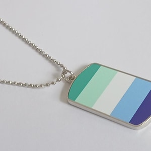 May include: A silver dog tag pendant on a ball chain necklace. The pendant has a rectangular shape with rounded edges and a design of horizontal stripes in shades of green, blue, and white. The pendant is a symbol of the gay community.