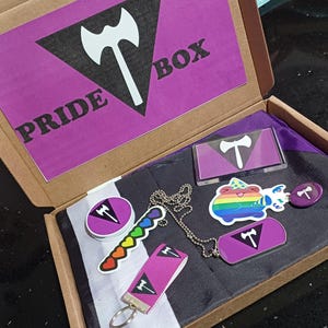 Puede incluir: Una caja morada y negra con el texto "PRIDE BOX" en la tapa. La caja contiene una variedad de artículos, incluyendo una bandera morada y negra, un collar con un colgante morado, una pegatina con un diseño de corazón arcoíris y un pequeño llavero morado y negro.