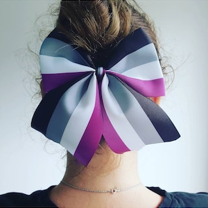 Puede incluir: Un gran lazo morado, blanco, negro y gris con un patrón de rayas. El lazo está atado en un nudo y se lleva en el pelo.