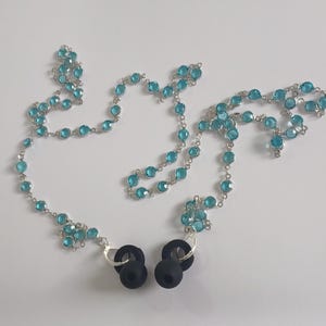 Può includere: Una catena d'argento ornata di perline sfaccettate rotonde di colore azzurro chiaro. La catena è collegata a due punte di auricolari nere. Le perline sono uniformemente distanziate, creando un design delicato. Gli auricolari sono neri e rotondi.