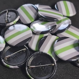 May include: A close-up of a pile of round, green, grey, and black striped buttons. The buttons have a pin back and are arranged in a random pattern.