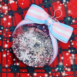 Może przedstawiać: Szklana bombka choinkowa w przezroczystym kolorze z wzorem śnieżynki z białego i srebrnego brokatu. Bombka jest ozdobiona niebieską, różową i białą kokardką.