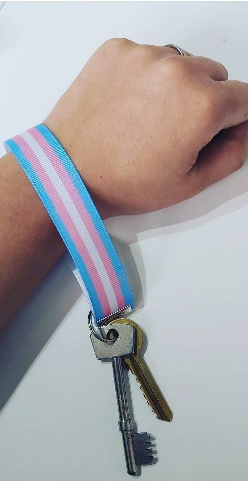 Puede incluir: Un cord&oacute;n de mu&ntilde;eca con los colores de la bandera del orgullo transg&eacute;nero: azul claro, rosa y blanco. El cord&oacute;n est&aacute; sujeto a un llavero plateado que sostiene dos llaves, una plateada y otra dorada.