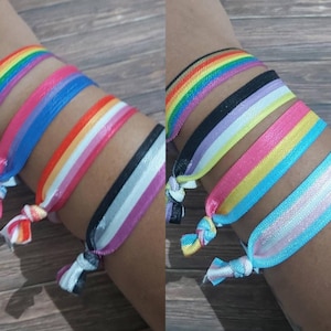 Op de afbeelding: Een set van zes kleurrijke elastische armbanden met verschillende regenboogvlagontwerpen. De armbanden zijn gemaakt van satijnen lint en zijn vastgebonden met een knoop. De armbanden worden om de pols gedragen.