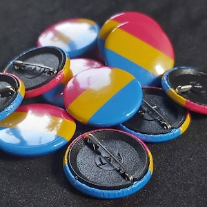 Puede incluir: Primer plano de un montón de botones redondos y coloridos con un diseño de bandera del orgullo pansexual. Los botones son azules, amarillos y rosas. Cada botón tiene una parte posterior con un alfiler.