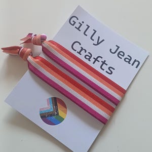 May include: Two hair ties with a striped design in shades of pink, orange, and white, resting on a white card. The card features the text "Gilly Jean Crafts" and a rainbow-colored logo.