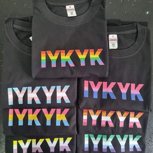 May include: Six black t-shirts with the text "IYKYK" printed in rainbow colours. Each shirt features a different variation of the rainbow flag.
