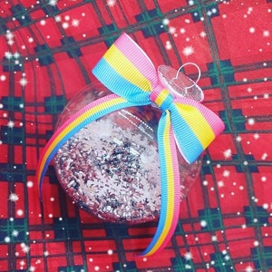 Op de afbeelding: Een transparante glazen kerstbal gevuld met zilveren glitter en versierd met een roze, blauw en geel lint dat in een strik is gebonden.