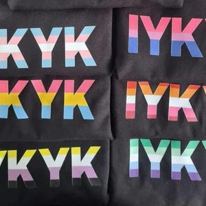 Könnte beinhalten: Fünf schwarze T-Shirts mit dem Text "IYKYK" in Regenbogenfarben bedruckt. Jedes Shirt hat ein anderes Farbschema, das verschiedene Pride-Flaggen repräsentiert.