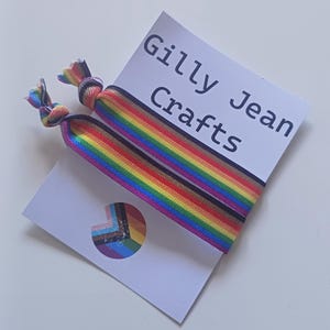 May include: Two rainbow-striped hair ties are shown on a white card with the text "Gilly Jean Crafts." A small circular sticker with a rainbow design is also visible. The hair ties are made of a colourful, elastic material.