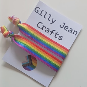 Peut inclure: Deux élastiques à cheveux rayés arc-en-ciel avec des extrémités nouées, posés sur une carte blanche imprimée avec "Gilly Jean Crafts". Les élastiques présentent un spectre de couleurs vives, dont le rouge, l'orange, le jaune, le vert, le bleu et le violet. Un petit autocollant aux couleurs de l'arc-en-ciel est visible.