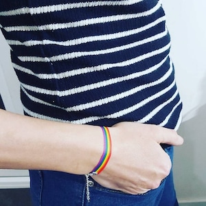 Könnte beinhalten: Ein Regenbogen-Armband mit einer silbernen Kette wird am Handgelenk getragen. Das Armband zeigt ein Farbspektrum, darunter Rot, Orange, Gelb, Grün, Blau und Lila. Die Person trägt ein marineblau-weiß gestreiftes Oberteil und blaue Jeans.