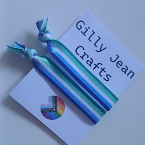 Pode incluir: Duas fitas de cabelo listradas em azul, verde e branco, apresentadas em um cartão branco. O cartão apresenta o texto "Gilly Jean Crafts" e um pequeno logotipo de arco-íris. As fitas são amarradas nas extremidades.