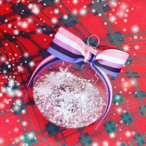 Op de afbeelding: Een transparante glazen kerstbal met zilveren glitter en witte sneeuwvlokken erin. De bal is versierd met een roze, wit, zwart en blauw lint dat in een strik is gebonden.
