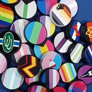 May include: A selection of circular phone grips, each featuring a unique design in vibrant colours. Designs include rainbow stripes, paw prints, and geometric patterns. The grips have black edges and are on a blue surface.