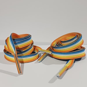 Pode incluir: Dois pares de cadarços coloridos, cada um enrolado em espiral. Os cadarços apresentam um padrão listrado de laranja, amarelo, azul e branco. As pontas dos cadarços são visíveis. O fundo é branco liso.