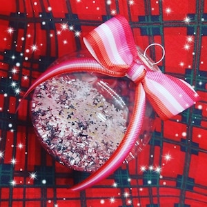 May include: A clear glass Christmas ornament with pink, white, and orange glitter inside. A pink, white, and orange ribbon is tied around the ornament in a bow.