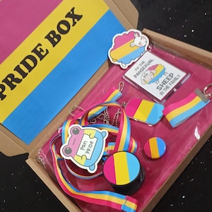 Könnte beinhalten: Eine Pride Box gefüllt mit pansexuellen Artikeln, darunter ein Lanyard, ein Schlüsselanhänger, ein Aufkleber, ein Knopf und eine Flagge. Die Box ist mit pinkfarbenem Stoff ausgekleidet und hat oben eine gelbe und blaue Flagge.