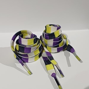 Puede incluir: Dos pares de cordones planos, enrollados en paquetes. Los cordones presentan un patrón repetitivo de cuadrados blancos, amarillos, negros y morados. Los terminales son plateados y amarillos. El fondo es blanco.