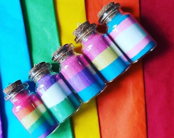 May include: Five small glass bottles with cork stoppers filled with colorful sand. The bottles are arranged in a row on a rainbow background. Each bottle contains a different color combination of sand, representing the colors of the pride flag.