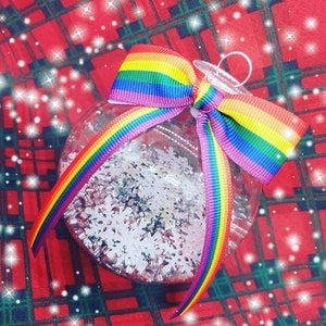 Op de afbeelding: Een transparante glazen kerstbal met een regenboog lint strik. De bal is gevuld met witte glitter en sneeuwvlokken.
