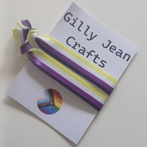 May include: Two non-binary pride flag hair ties with yellow, white, purple, and black stripes. The hair ties are on a white card with the text "Gilly Jean Crafts" and a rainbow pride flag sticker.