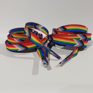 Pode incluir: Um par de atacadores planos com um esquema de cores do arco-íris, com um design de bandeira do orgulho progressista. Os atacadores são vermelhos, laranja, amarelo, verde, azul e roxo, com detalhes brancos, rosa, azul claro, preto e castanho.
