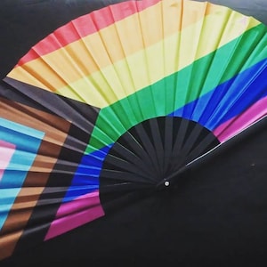 May include: A rainbow-colored folding fan with a black handle. The fan features the colors of the rainbow pride flag, as well as the colors of the transgender pride flag.