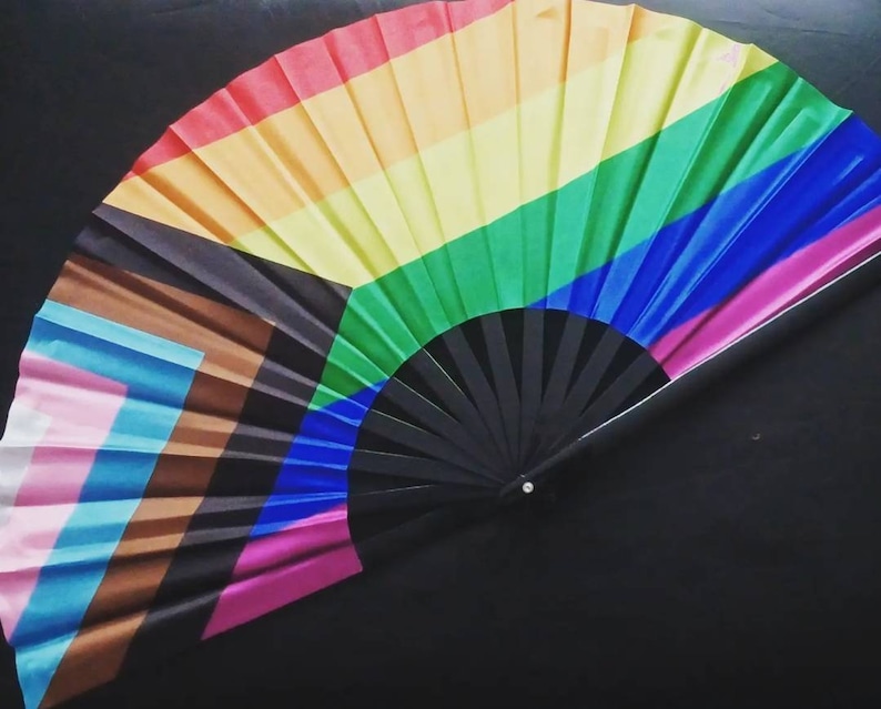 May include: A rainbow-colored folding fan with a black handle. The fan features the colors of the rainbow pride flag, as well as the colors of the transgender pride flag.