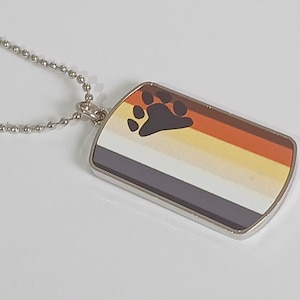 May include: A silver dog tag pendant on a ball chain necklace. The tag features the Bear Pride flag design with a paw print in the upper left corner. The flag has stripes of brown, orange, yellow, white, gray, and black.