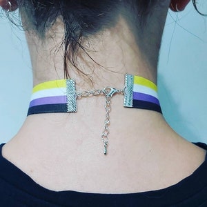 Puede incluir: Un collar gargantilla negro con una cinta amarilla, blanca, morada y negra. La cinta está en los colores de la bandera del orgullo no binario. El collar tiene una cadena y un cierre plateados.
