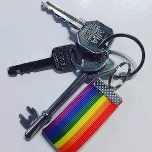 Könnte beinhalten: Ein Schlüsselsatz mit einem Schlüsselband aus Regenbogenband. Die Schlüssel sind silberfarben und haben verschiedene Formen und Größen. Das Band ist aus weichem, buntem Stoff und oben zu einem Knoten gebunden.