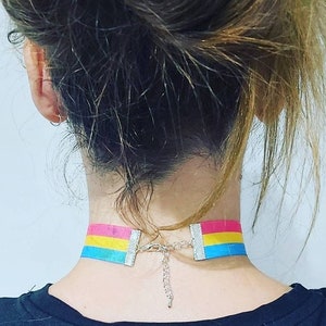 May include: A person wearing a choker necklace with a pansexual pride flag design. The choker is made of blue, yellow, and pink ribbon with a silver chain closure.
