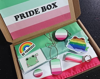 May include: A green, pink, and white pride flag is in a box with a variety of pride-themed items, including a frog keychain, a necklace with a tag, a sticker, and a pin. The box is labeled "PRIDE BOX" and the sticker says "BE YOURSELF".