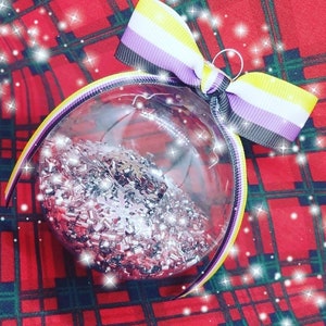 Może przedstawiać: Szklana, przezroczysta ozdoba choinkowa wypełniona srebrnym brokatem. Ozdoba jest ozdobiona fioletową, żółtą i czarną wstążką zawiązaną w kokardkę.