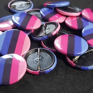 May include: A collection of 1.25 inch round pinback buttons with a bi pride flag design. The buttons are blue, pink, and black with a white pinback.