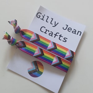 Peut inclure: Deux élastiques à cheveux colorés avec un motif en chevrons arc-en-ciel et une étiquette indiquant "Gilly Jean Crafts". Les élastiques ont un nœud à motifs arc-en-ciel et rose, bleu et blanc.