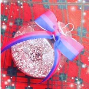 Op de afbeelding: Een transparante glazen kerstbal met zilveren glitter erin. Hij is versierd met een blauw, roze en wit lint dat in een strik is gebonden.