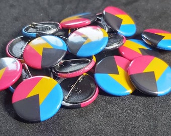 May include: A collection of round pinback buttons with a colorful geometric design. Each button features a segmented pattern in pink, yellow, black, and blue. The pins are scattered, showing the pin backs.
