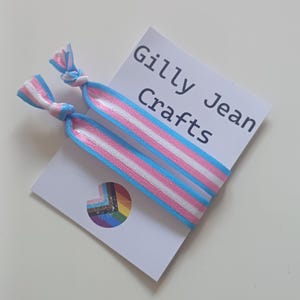 Könnte beinhalten: Zwei Haargummis mit blauem, weißen und rosa Streifenmuster, die die Transgender-Pride-Flagge darstellen. Die Gummis sind an den Enden verknotet. Eine weiße Karte mit dem Text "Gilly Jean Crafts" befindet sich hinter den Gummis.