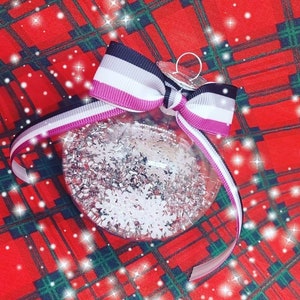 May include: A clear glass Christmas ornament with silver glitter and white snowflakes inside. It is decorated with a black, white, and purple ribbon tied in a bow.
