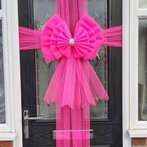 May include: A bright pink tulle bow with a silver accent is tied around a black front door. The tulle is draped down the sides of the door.