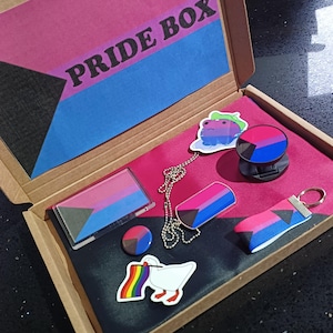 May include: A cardboard box with the words "PRIDE BOX" on the lid. The box is open and contains a variety of items, including a necklace, a keychain, a phone grip, a sticker, and a small makeup palette. All of the items are decorated with the bisexual pride flag.