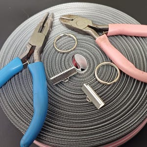May include: Two pairs of pliers, one blue and one pink, are lying on a roll of black and white striped ribbon. Two silver metal clasps are also on the ribbon.